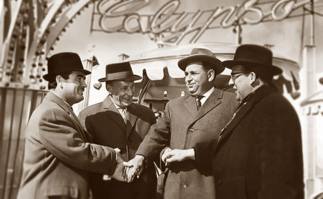 Vier Männer im Anzug stehen einander zugewandt vor einem Fahrgeschäft und geben sich die Hände. Das Bild ist in Schwarz-Weiß.