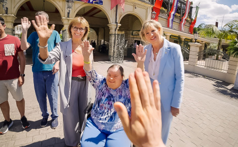 Mehrere Gäste stehen im Bereich des Haupteingangs vom Europa-Park und winken in die Kamera. Eine Person sitzt im Rollstuhl und hebt beide Hände fröhlich in die Luft.